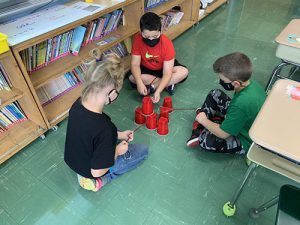 Three third-grade students sit on the floor with a pyramid of red cups. They are using strings to move the cups into place.