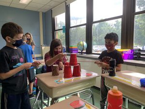 Three Young students use strings to stack red plastic cups on a desk.