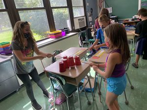 Three third grade students stand around desks. One has five plastic cups on it. The girls are holding string attached to the cups.