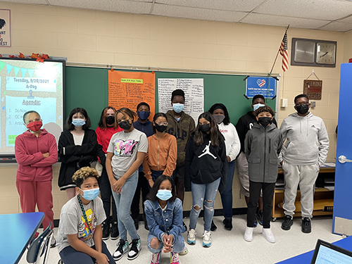 A class of 14 middle school students stands together in front of a blackboard with posters hanging from the top. All have masks on. Twelveof them are standing along the wall and two are kneeling in front.