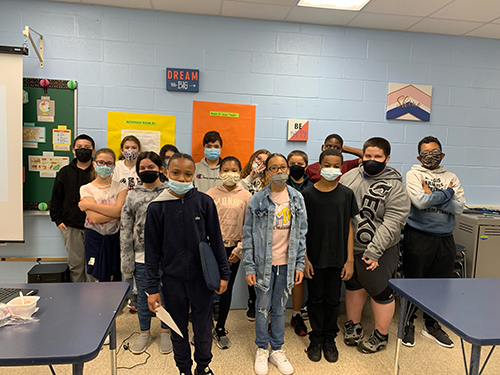 Fifteen middle school sixth grade students stand in three rows, in front of a light blue wall. All have masks on.