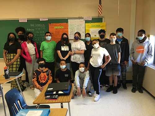 Fifteen eighth grade students stand in a classroom. They are in two rows, with three kids sitting on the floor and the rest standing behind them. All are wearing masks.