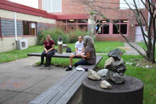 Outdoors, two high school students and a female teacher sit on benches. There is a tree in the background with windchimes on it and rocks piled on top of a light structure. There is grass behind them and a patio in front.