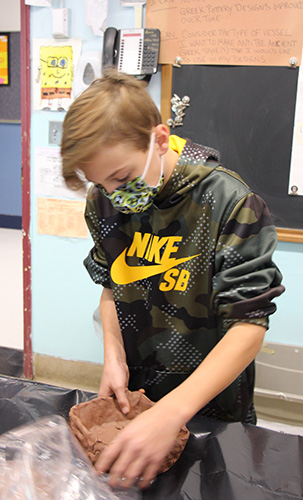 A middle-school age boy, with short light hair and wearing a camouflage Nike sweatshirt and green mask, molds clay into a bowl.