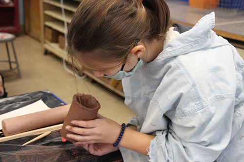 A sixth-grade girl with long brown hair in a ponytail, wearing a blue smock and blue mask, looks into a cup she is making out of clay.