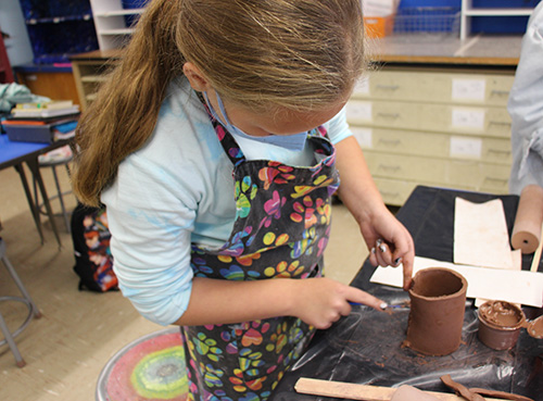 A sixth-grade girl with long blonde hair in a ponytail, wearing a multi-colored apron, uses a knife on the clay she is shaping into a cup.