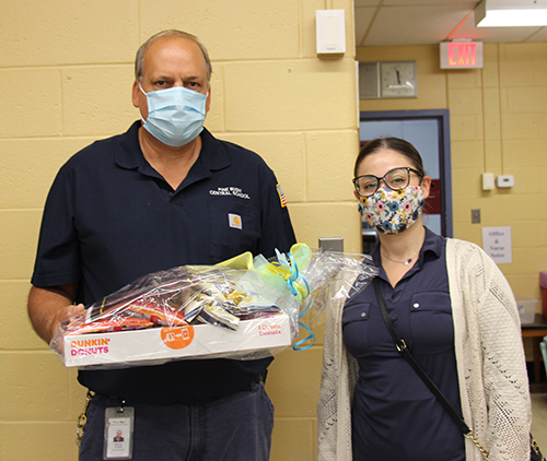 A man in a dark blue workshirt that says Pine Bush Central Schools and a light blue mask holds a package of donuts and snacks. On his right is a woman in glasses, blue shirt and tan sweater.