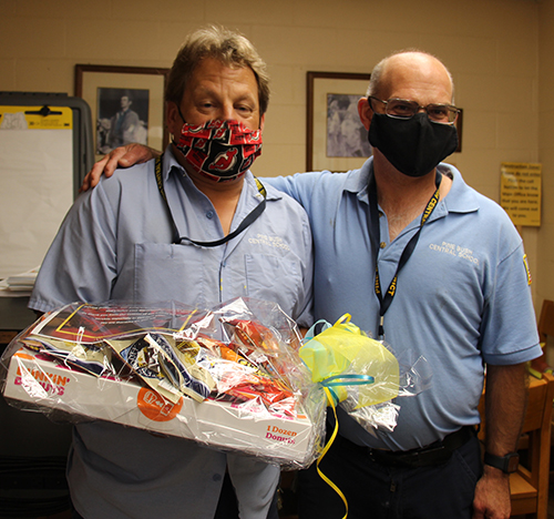 Two men, both wearing light blue shirts and masks stand togeher