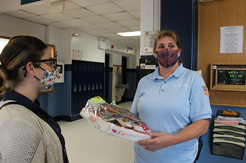 A woman in a light blue polo shirt and dark mask holds a package with donuts and snacks. On her left is a woman in a tan sweater and mask looking at her.