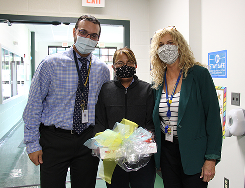 On the left is a man in a blue dress shirt and tie, wearing a mask, in the center is a shorter woman with navy blue shirt and mask holding a gift of a box of donuts and snacks, on the right is a woman in a blue and white striped shirt and blue sweater, light colored mask.