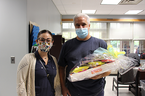 A woman wearing a blue shirt and tan sweater, glasses and a flowered mask stands next to a man in a blue shirt and mask. He is holding a gift bag with donuts and snacks in them.