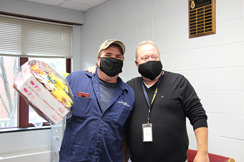 A man wearing a blue shirt and a baseball cap and black mask holds a package with donuts and snacks. Next to him is another man wearing a black shirt and mask.