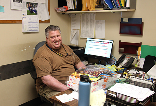 A man in a brown shirt, with salt and pepper hair, sits at a desk with a computer to one side. There are two boxes of donuts and snacks wrapped in cellophane on the desk. He is smiling.