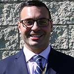 A man with short dark hair, wearing glasses, a blue suit jacket, white shirt and blue and gold striped tie, smiles. There is a block wall behind him.