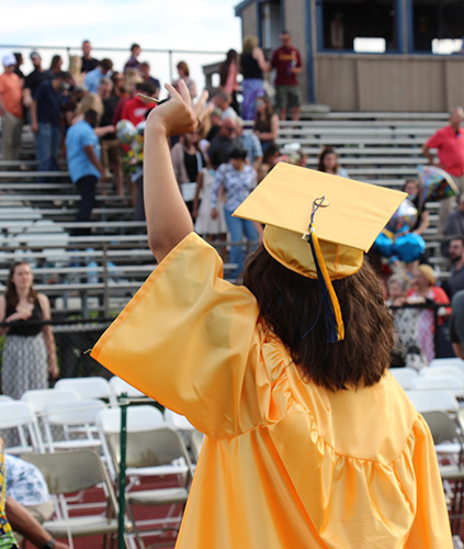 A young woman with shorter dark hair has her back to the camera is wearing a graduation cap and gown. She is waving to people in the stands.