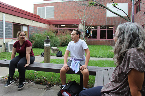 Two high school students sit on a bench with a teacher sitting on another bench. They are outdoors, with grass and a tree in the background. 