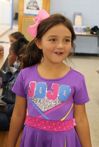 An elementary age girl with long dark hair wearing a purple dress, with the word JOJO on it in pink and blue. She has a big pink bow in her hair.