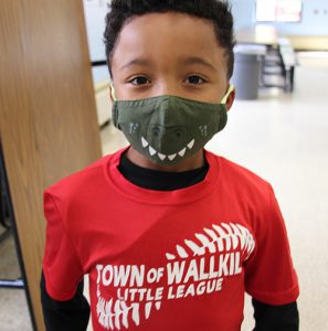 A little boy wearing a green mask with smiling teeth has on a red Town of Wallkill Little LEague shirt on. He has short dark hair.
