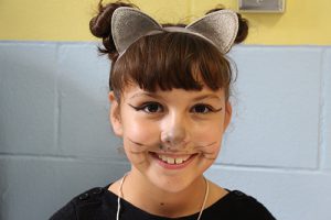 An elementary age girl with dark hair and bangs. She has her face panted with whiskers like a cat and a headband with cat ears. 