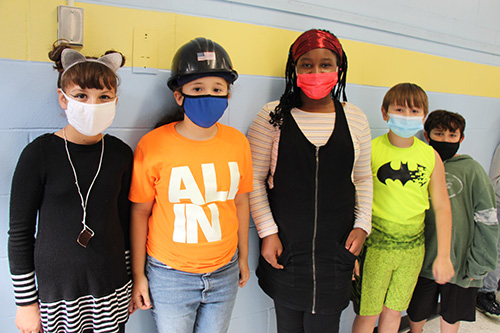 Five elementary students stand together, all wearing masks. They are wearing clothes to show their interests - one is a cat, an electrician, batman, a jets fan.