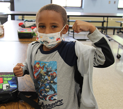 An elementary age boy makes a muscle with his arm. He is wearing a gray shirt with the Avengers cartoon characters on it. He alsohas a white mask with different color characters on it.