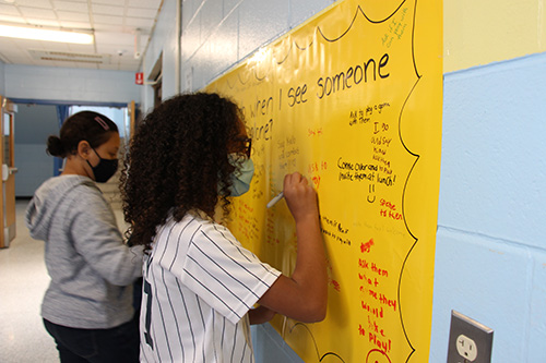 An older elementary age girl, with shoulder length curly dark hair and wearing a yankees jersey, writes on a large yellow poster. There are two other kids beside her also writing on it.