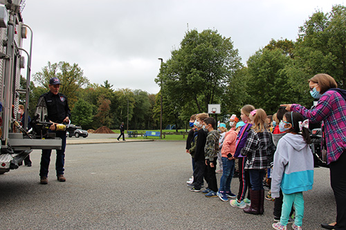 A group of elementary school kids stand to the right as a firefighter, dressed n a blue uniform holds a piece of equipment from his fire truck. 