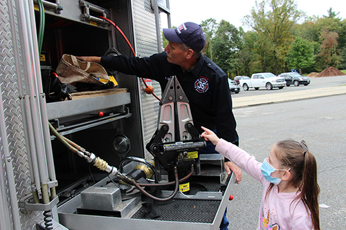 A girl wearing a pink shirt and blue mask points into a fire truck. The firefighter looks in. He is wearing a blue uniform and hat.