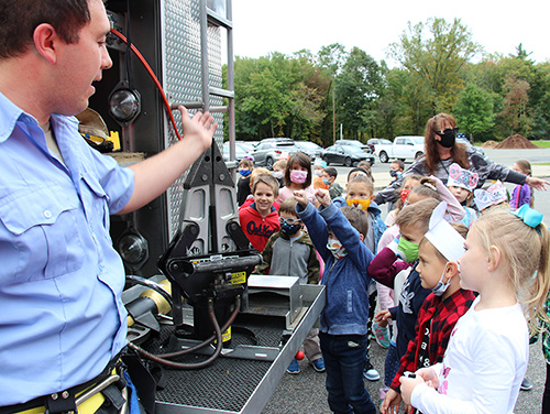 A firefighter wearing a light blue shirt and dark blue pants holds his hand out toward his truck and equipment. A group of elementary school kids are gathered around watching. One boy in front holds his arms up in victory after correctly answering what type of equipment it is.