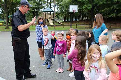 A volunteer firefighter, wearing dark blue pants and shirt and hat, talks to a group of elementary age students.