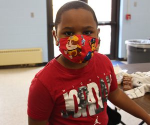 An elementary age boy wears a red shirt with the word JORDAN on the front and pictures of Michael Jordan on it. He has a red mask on.