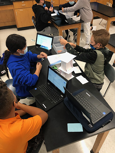 Looking down onto a lab table where three sixth-grade boys are working. There are chromebooks open and a tower of index cards in the center.
