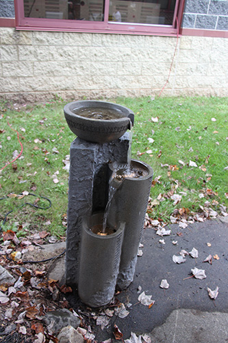 A small bowl with a spout sits on top of a concrete column. Water falls from the gowl down to another bowl and from that bowl to another to make a fountain.