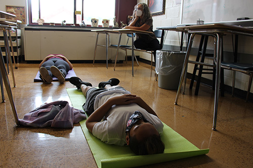 Students lay on the floor on yoga mats with a teacher sitting at a desk in the background. They are all masked.