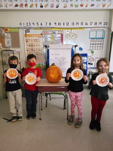 Four kindergarten kids, two boys on the left and two girls on the right -  hold up paper plates they made into pumpkins. In between them is a jack-o-lantern.