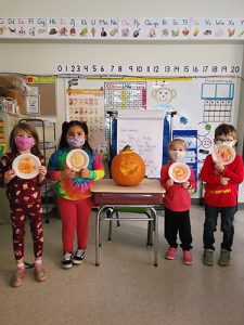 Four kindergarten students hold plates up with their drawn pumpkins on them. In the center of them is a large jack-o-lantern.