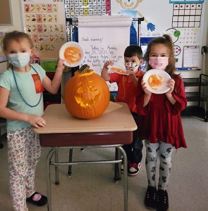Two little girls in front and a boy in back hold up paper plates with orange on them. On a desk between them is a large jack-o-lantern.