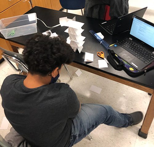 A sixth-grade boy sits at a black lab table looking at a tower of index cards. There is an open Chromebook on the table.