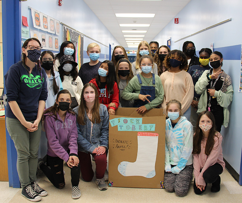 A large group of middle-school age kids stand together with a large brown box in front of them. On the box is a large cut out of a sock. All kids have masks on.