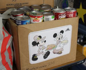 A large brown cardboard box with many cans stacked neatly inside. Outside is a drawing of mickey and Minnie Mouse dressed as pilgrims holding turkey and pie.