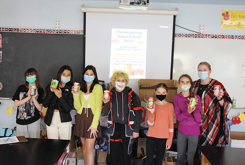 Six middle school students stand in front of boxes and boxes of collected canned goods. Each is holding a can in their hand. On the right is their teacher, a woman, also holding a can. They all have masks on.