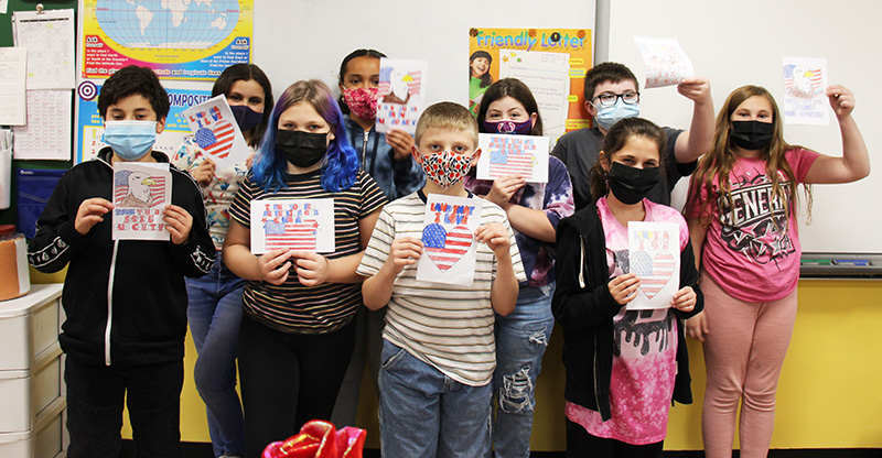 A group of nine fifth-grade students stand together holding up the cards they made for veterans. Most cards are red, white and blue. All kids are wearing masks.