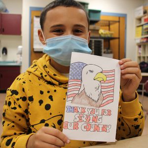A fifth-grade boy with short brown hair, blue mask and yellow hoodie with lack spots holds up his card he created for veterans day. It has a flag on it, bald eagle and the words Thank you for serving our country in red white and blue.