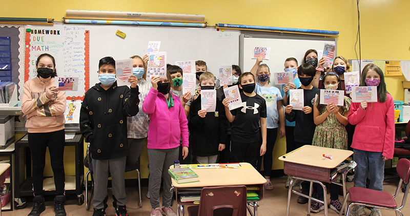 A class of fifth-grade students, about 20 of them, stand in front of a classroom holding up the cards they made for veterans. All students are masked.
