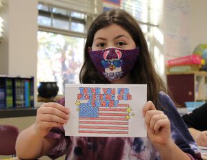 A girl with shoulder length brown hair holds up a card she made with a US flag on the cover and the words Thank you for your service to our country in red white and blue