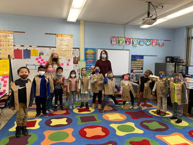 Twelve kindergarten students stand around a classroom, all wearing brown paper vests with a letter of the alphabet on it. There are two adults in the background. All are wearing face masks. They are standing on a multicolored rub with different shapes on it.