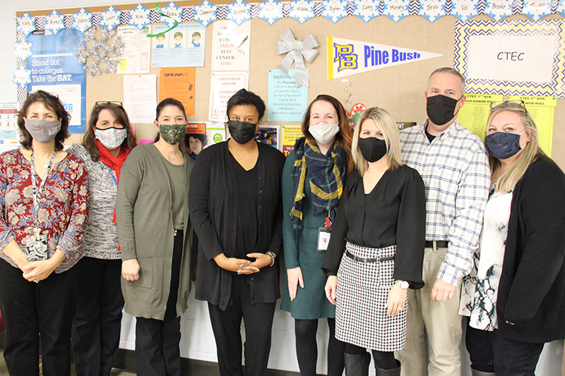A group of eight adults stand together in front of a bulletin board. All have masks. All are women except for one, second from right. In the background is a pennant that says Pine Bush.