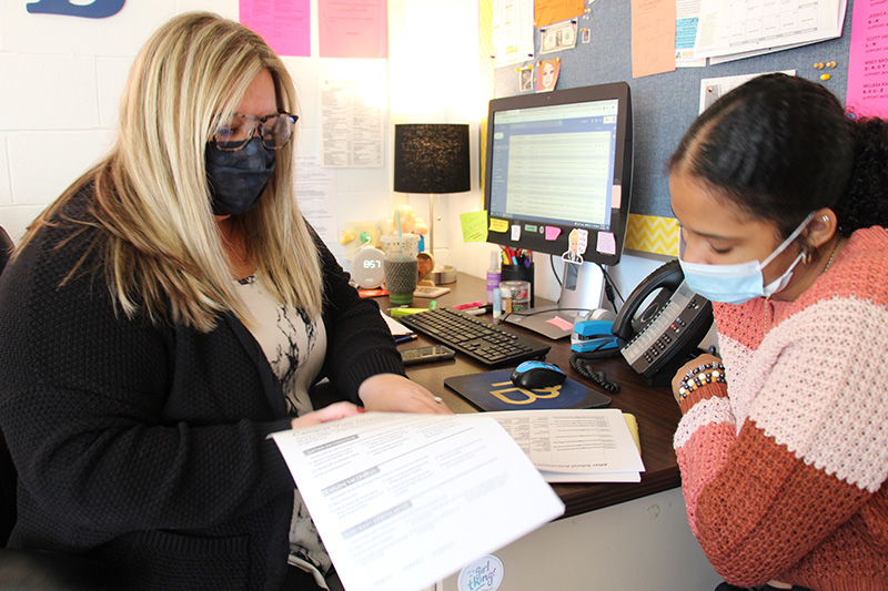 A woman sits on the left. She has blonde hair, is wearing a sweater, dark mask and glasses. She is holding papers and sitting at a desk talking to a high school student, wearing a blue mask and orange strip sweater. She has dark hair pulled back. 