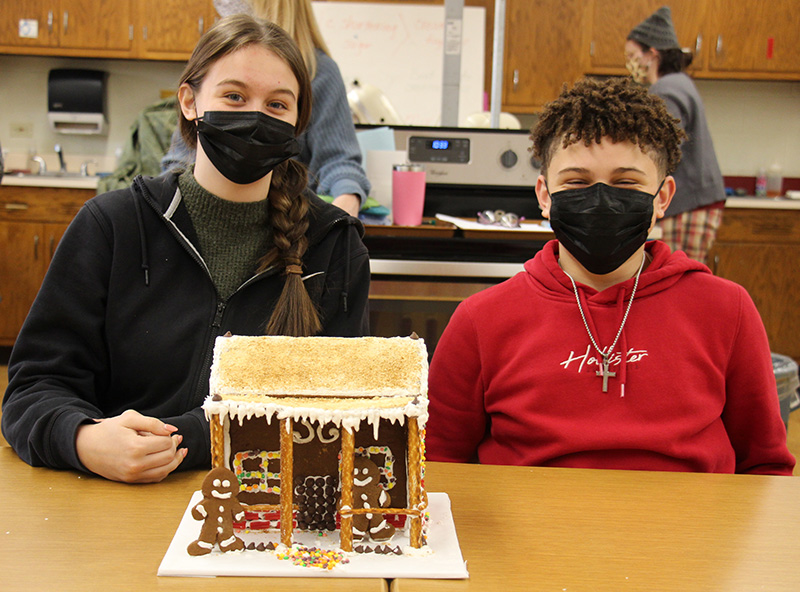 Two high school students sit at a table with a gingerbread house in front of them. They both are wearing black masks. The house has a brown roof and pretzels making a front porch.