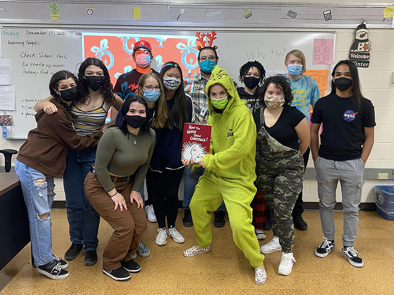 A class of 10 high school students stand together with a male teacher in back and another teacher in front dressed in a green one-piece grinch costume. She is holding a copy of the book "The Grinch Who Stole Christmas."